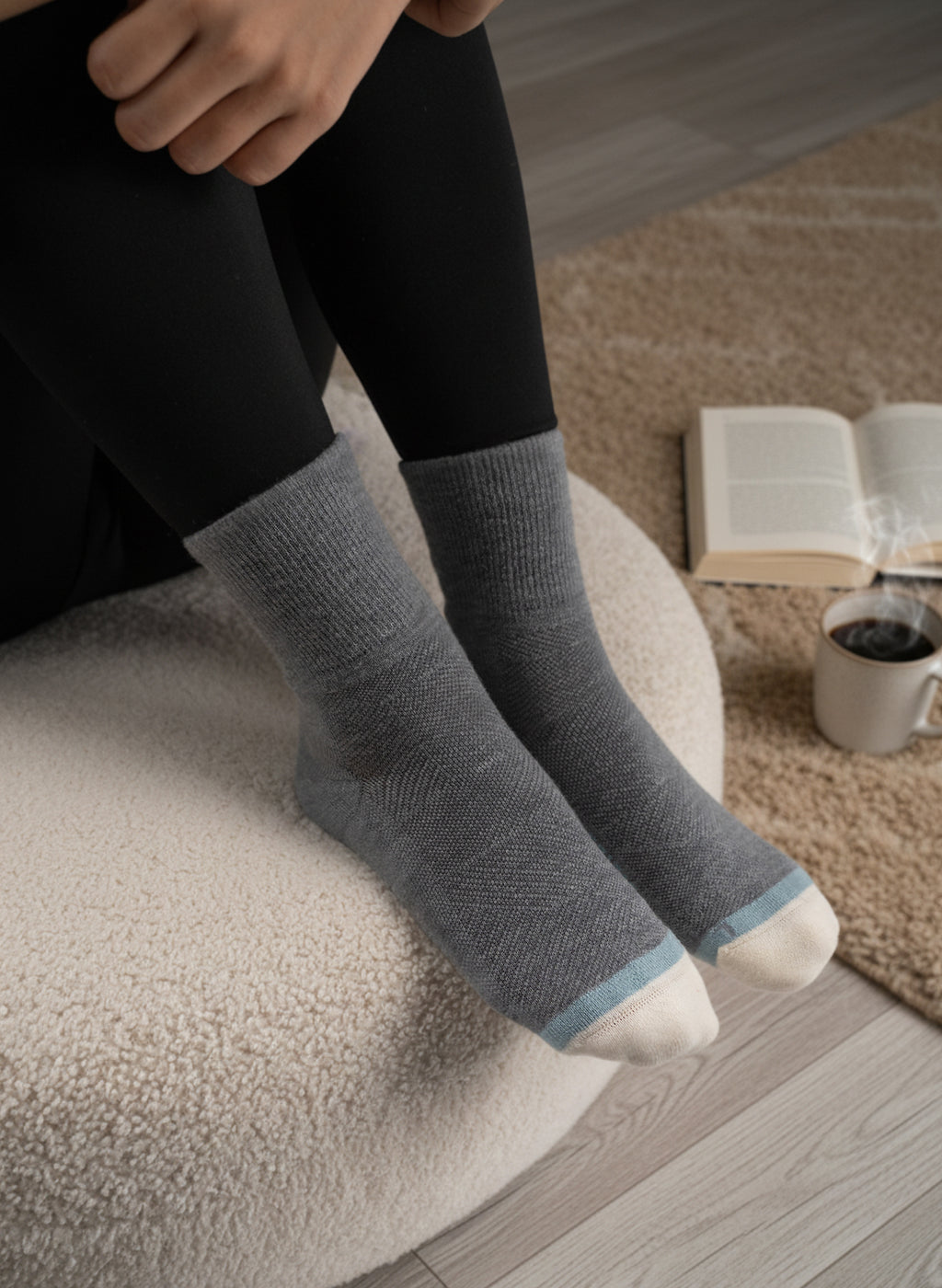 Person wearing gray EFISIO Merino Wool Crew socks with a blue accent on a cushioned chair, with a book and coffee cup in the background.